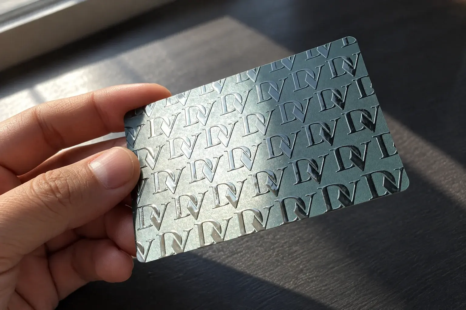 PVC vizit kartica sa metalik monogramom nagnuta ka prozoru, pruga svjetla preko uzorka
